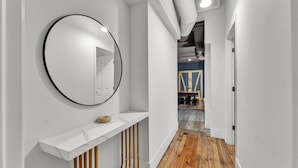 A hallway with a white wall, a round mirror, a marble shelf, wooden flooring, and a door leading to another room. - Apartmint at Arch St Philadelphia (Philadelphia)
