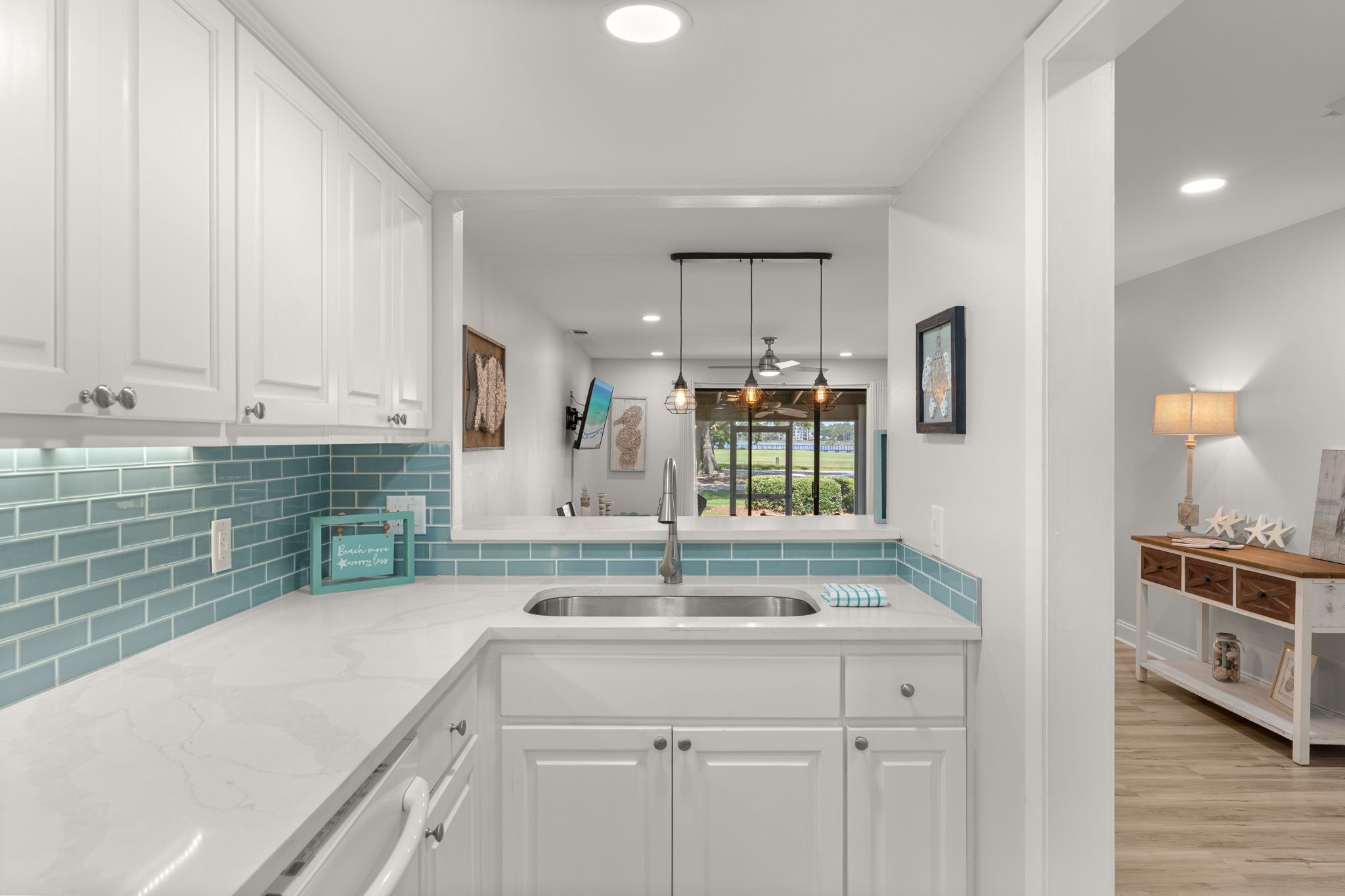 A kitchen with white cabinets, a blue-tiled backsplash, a stainless steel sink, pendant lights, and a view through a window.