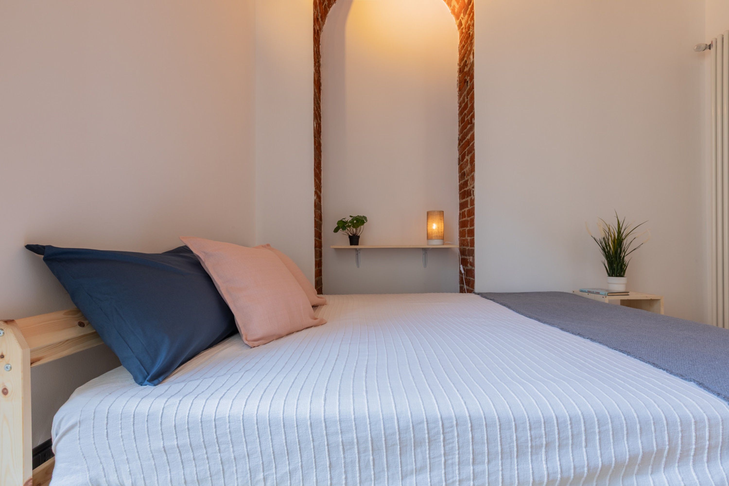 A bedroom with a bed, pillows, a nightstand, a wall-mounted shelf with a plant, and a wall-mounted light.