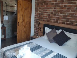 Ein Hotelzimmer mit Backsteinwand, einer Holztür, einem Bett mit Kissen, einem Nachttisch und einem durchgegangenen Tür erkennbaren Badezimmer.
