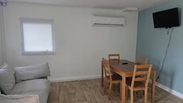 Un salon comprenant une table à manger en bois, un canapé, un appareil de climatisation, un téléviseur fixé au mur et une fenêtre avec des stores.