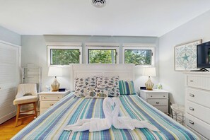 Ein Schlafzimmer mit einem Bett, zwei Nachttischen, einem Sessel, einem Fernseher und drei Fenstern.