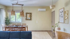 A living room with a blue sofa, a beige sofa, a dining table with chairs, a hanging rattan chandelier, an air conditioner, and wall-mounted artwork. - Ambela House, Traditional Living in Kefalos, Kos (Kos)