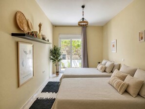 A bedroom with two beds, a large window with curtains, a hanging light fixture, and a framed picture on the wall.