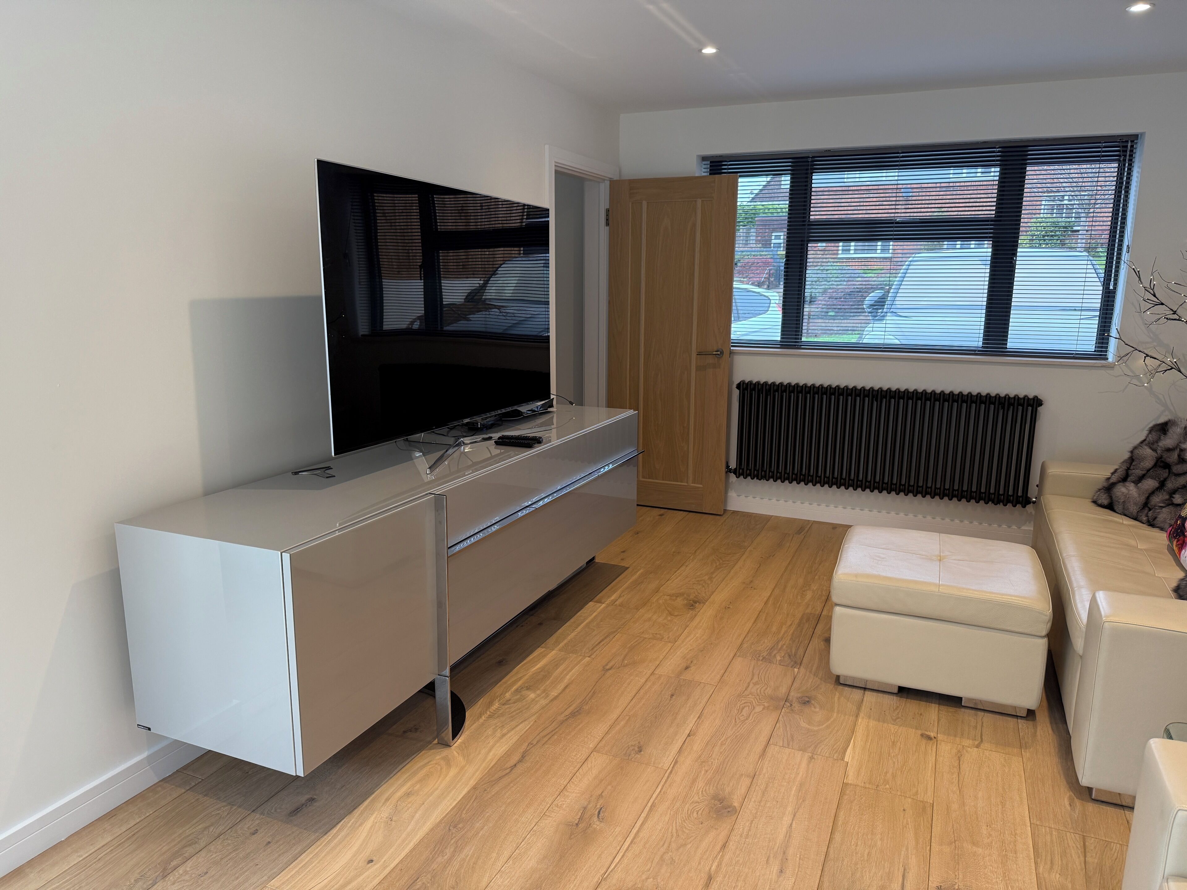 En moderne stue med et fladskærms-tv, en hvid sofa, en lille skammel og trægulv.