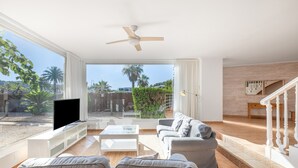 Una sala de estar con televisor, sofás, una mesa de centro, un ventilador de techo y vistas al exterior.