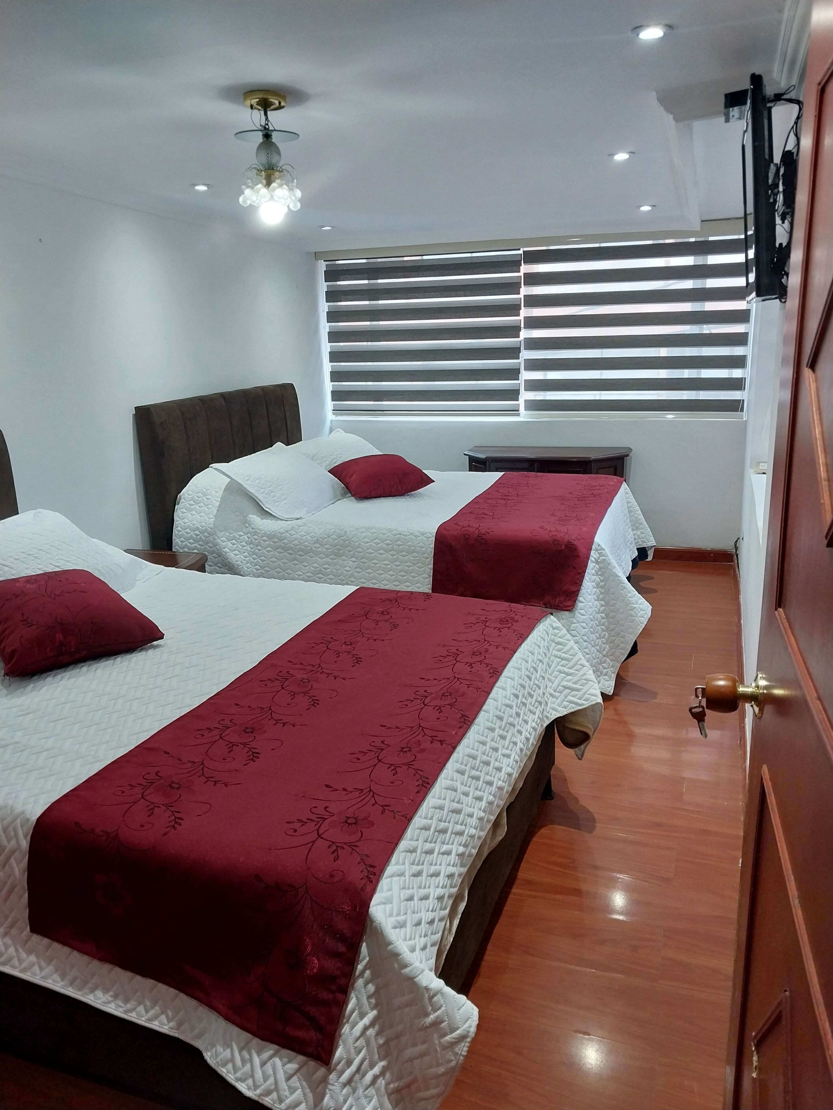A hotel room with two beds, a wooden floor, a window with blinds, and a ceiling light fixture.