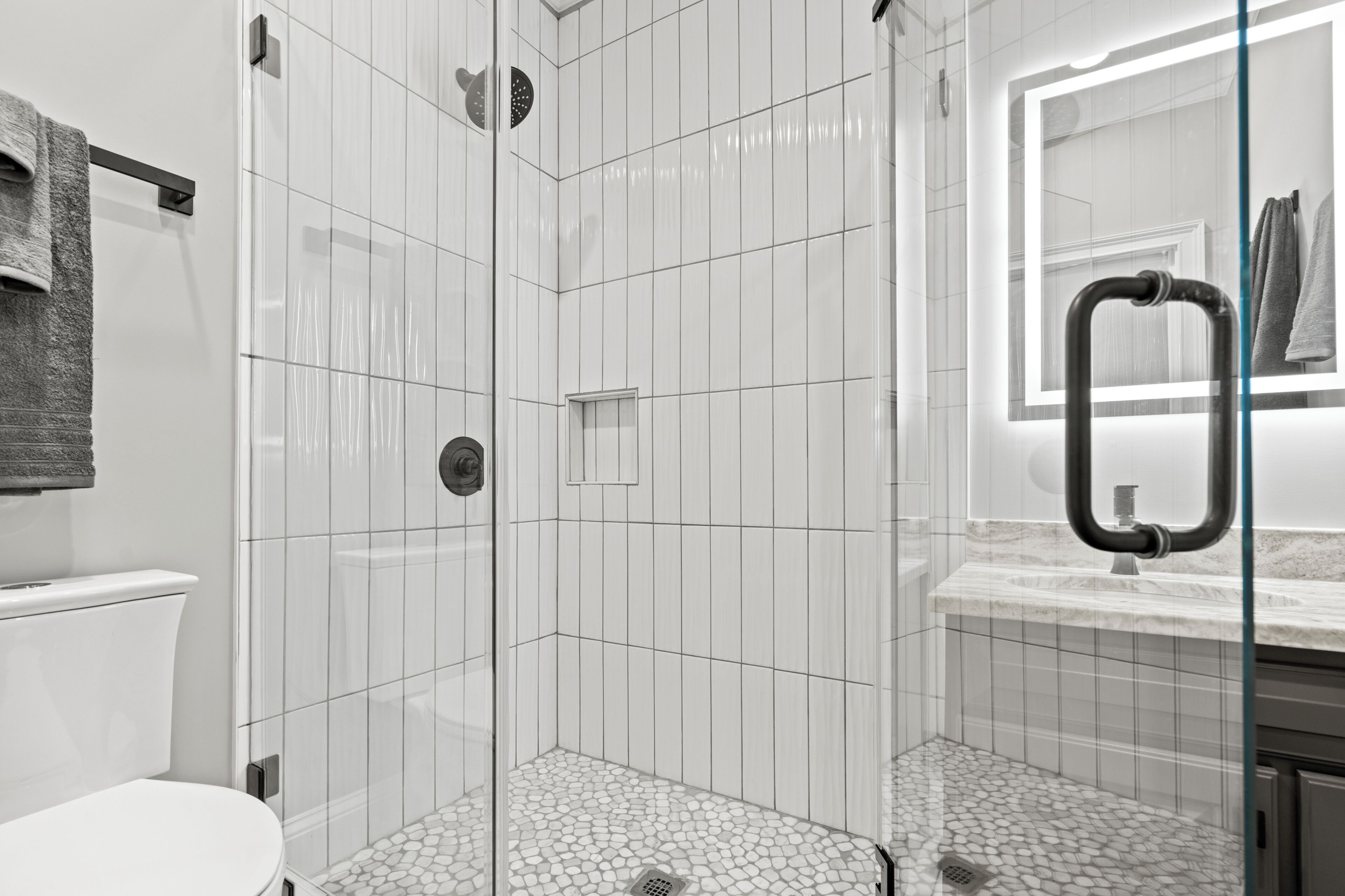 A modern bathroom with a glass-enclosed shower, white tiled walls, a marble countertop, and a visible toilet.