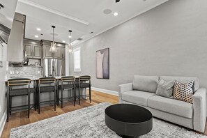 A modern kitchen with stainless steel appliances, a dining area with a table and chairs, and a living room with a sofa and a round black ottoman. - The Urban Flats (Florence)