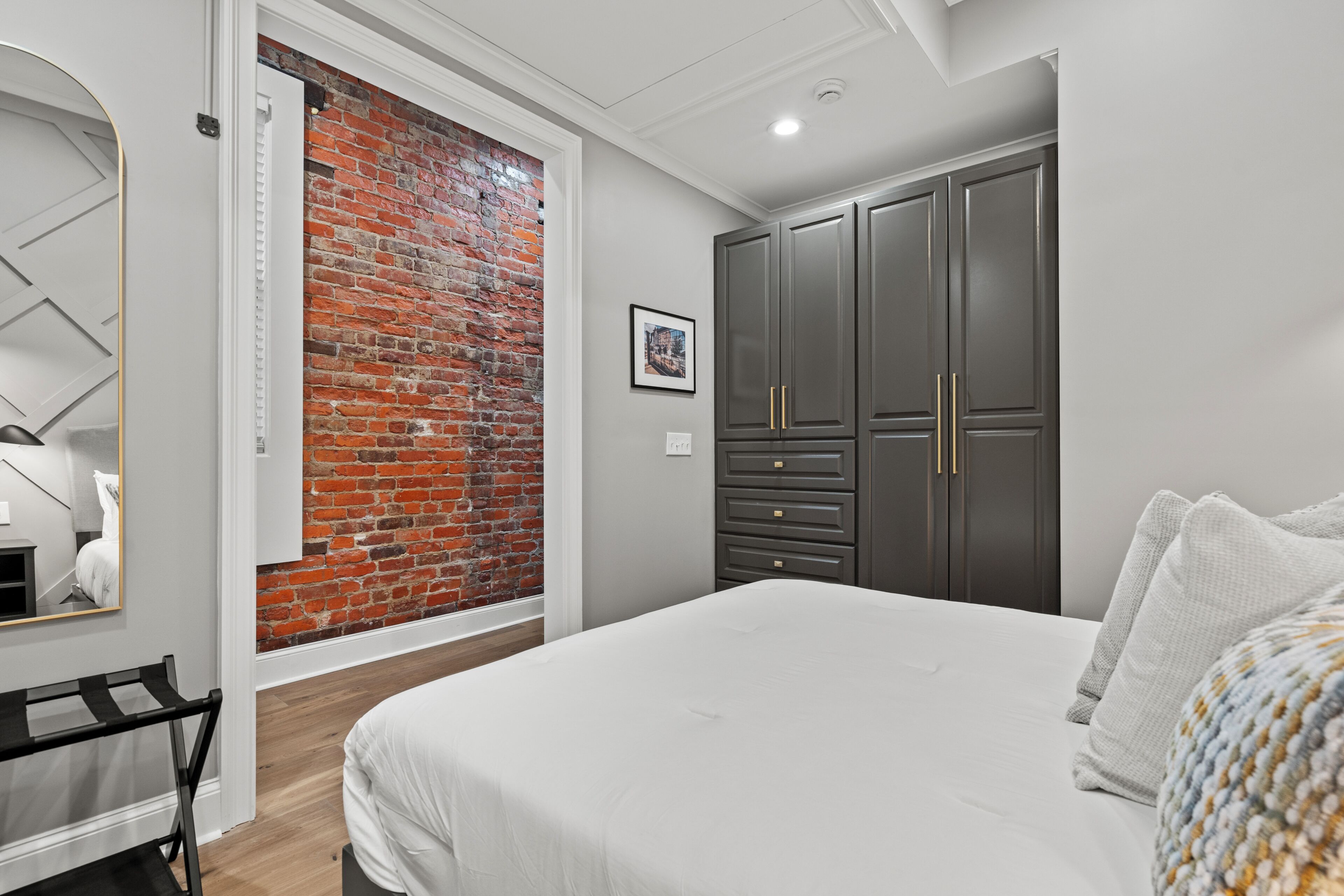 A bedroom with a brick wall, a bed with white bedding, a wardrobe with dark doors, and a mirror.