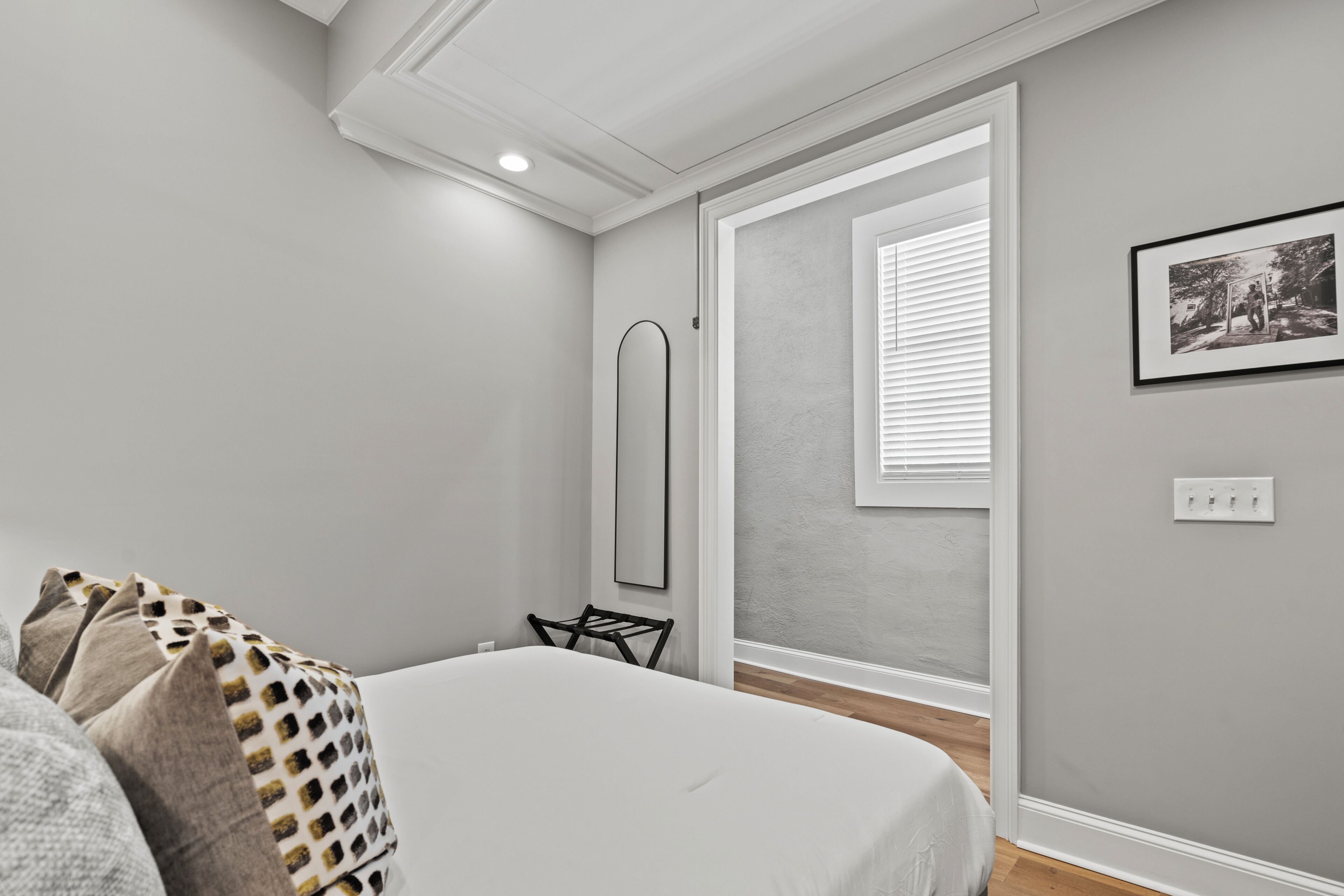Un dormitorio con cama, una silla, una ventana con persianas, un cuadro en la pared y un interruptor de luz.