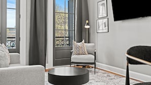 Uma sala de estar moderna com um sofá cinza, uma mesa de centro redonda, uma poltrona cinza com almofadas estampadas, um abajur e uma televisão grande fixada na parede.