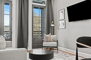 A modern living room with a grey sofa, a round coffee table, a grey armchair with patterned cushions, a floor lamp, and a large TV mounted on the wall.