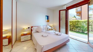A modern bedroom with a bed, bedside tables, a nightstand, and a view of greenery through a glass door. - Freestyle Apartment By Bookinggardalake (Salò)