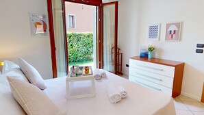 Une chambre d’hôtel avec un lit, une table de chevet sur laquelle se trouve une petite table avec des articles de toilette, un bureau avec une chaise, et une vue sur un jardin par une porte ouverte.