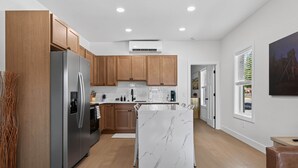 A modern kitchen with wooden cabinets, a stainless steel refrigerator, a marble island, and a view of the outdoors through a window.