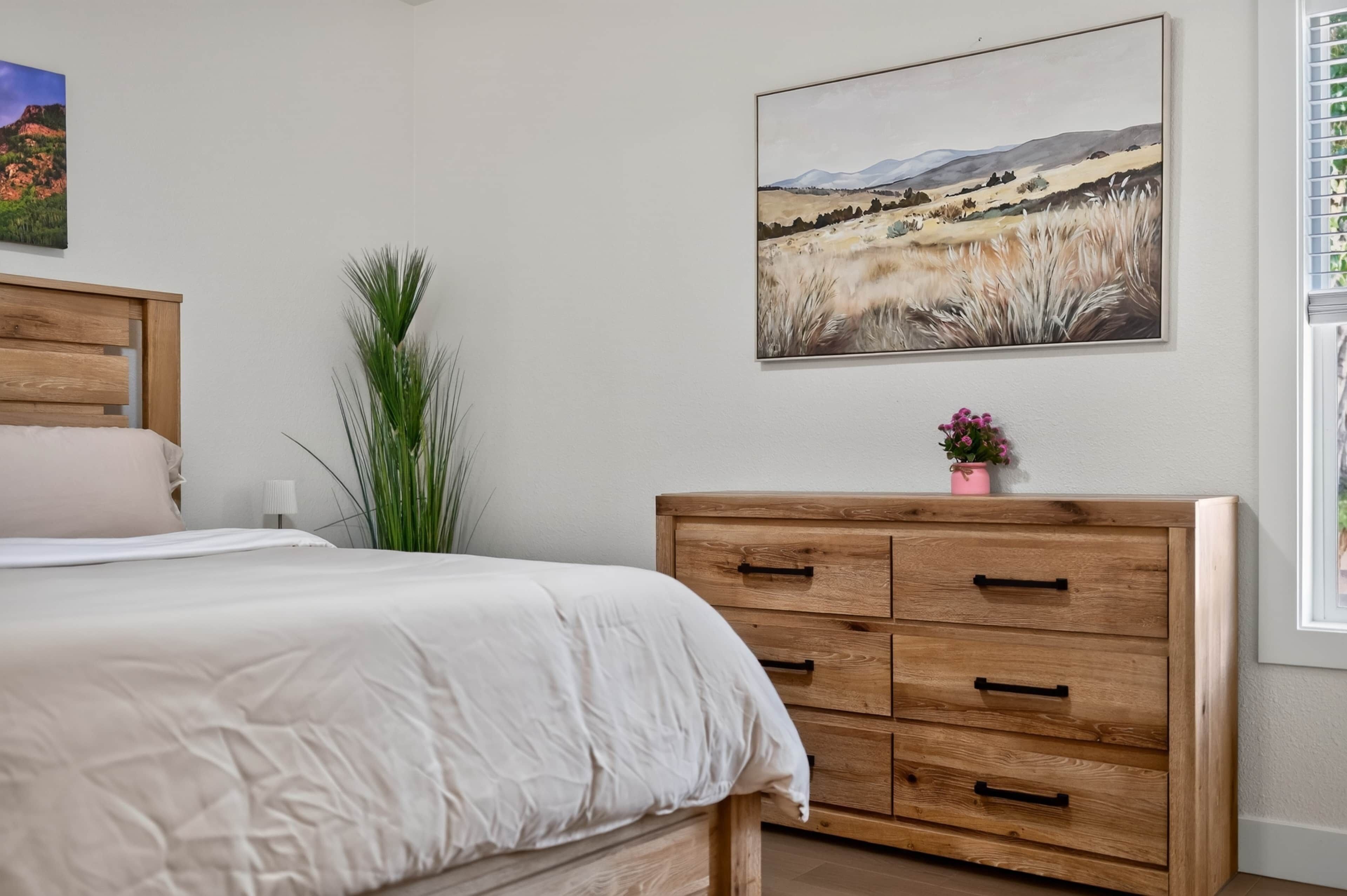 A bedroom with a bed, a wooden dresser, a landscape painting, and a potted plant.