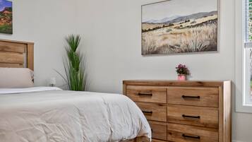 A bedroom with a bed, a wooden dresser, a landscape painting, and a potted plant.