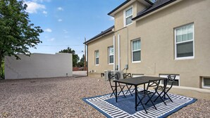 Un'area patio con tavolo e sedie, un tappeto azzurro e bianco e un edificio con diverse finestre e facciata beige.