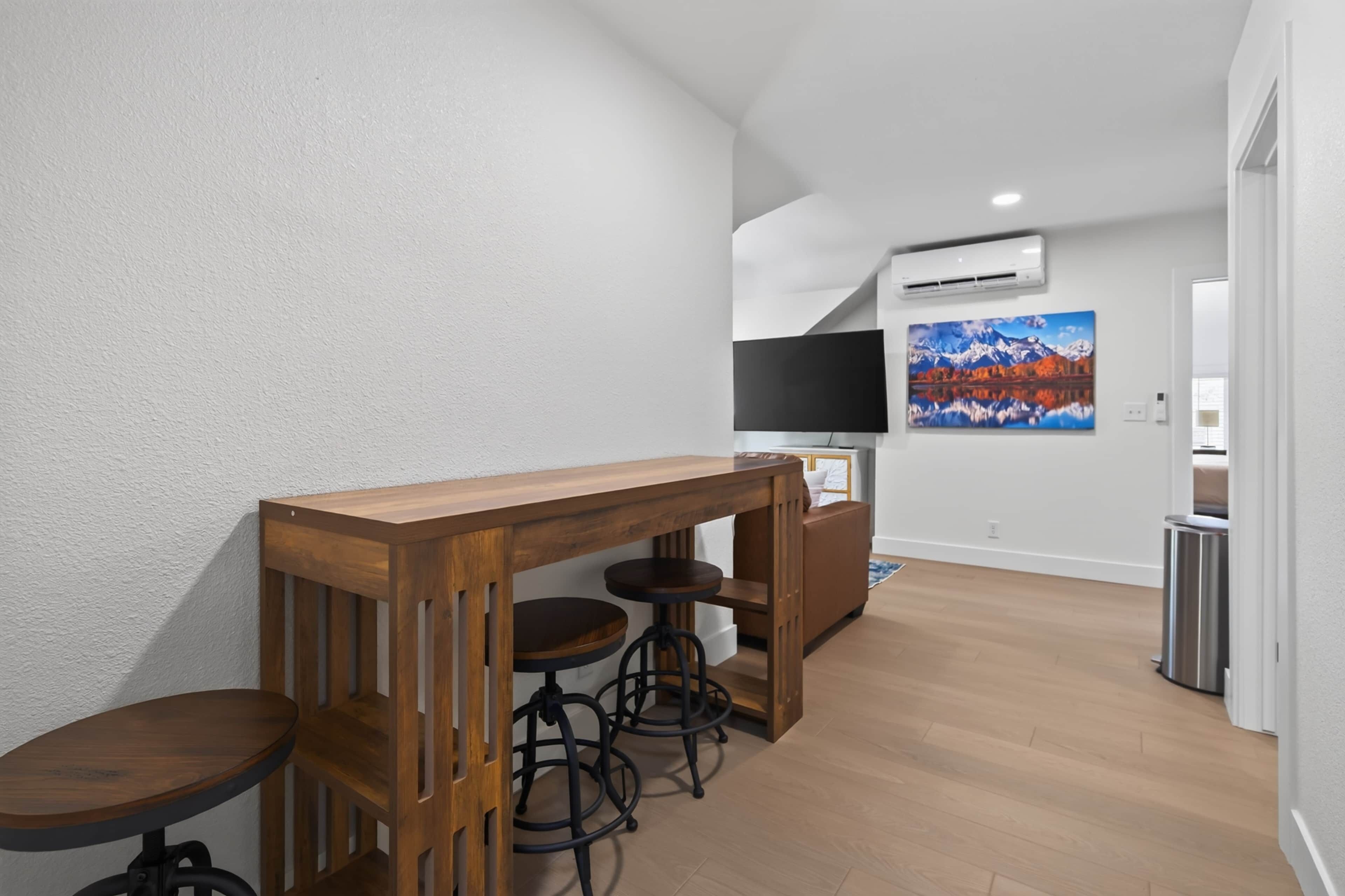 A modern kitchen with a wooden bar and stools, a flat-screen TV, and a painting of a mountain landscape.