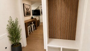 Un pasillo con una planta en maceta, suelo de madera, pared blanca con un cuadro y una zona de cocina al fondo.