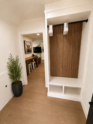 A hallway with a potted plant, a wooden floor, a white wall with a picture, and a kitchen area in the background.