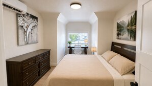 Un dormitorio con cama, cómoda, escritorio, silla, lámpara y dos cuadros.
