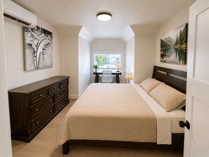 A bedroom with a bed, dresser, desk, chair, lamp, and two paintings.