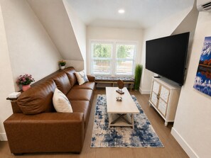 A living room with a brown leather sofa, a coffee table, a flat-screen TV, and a window with blinds.