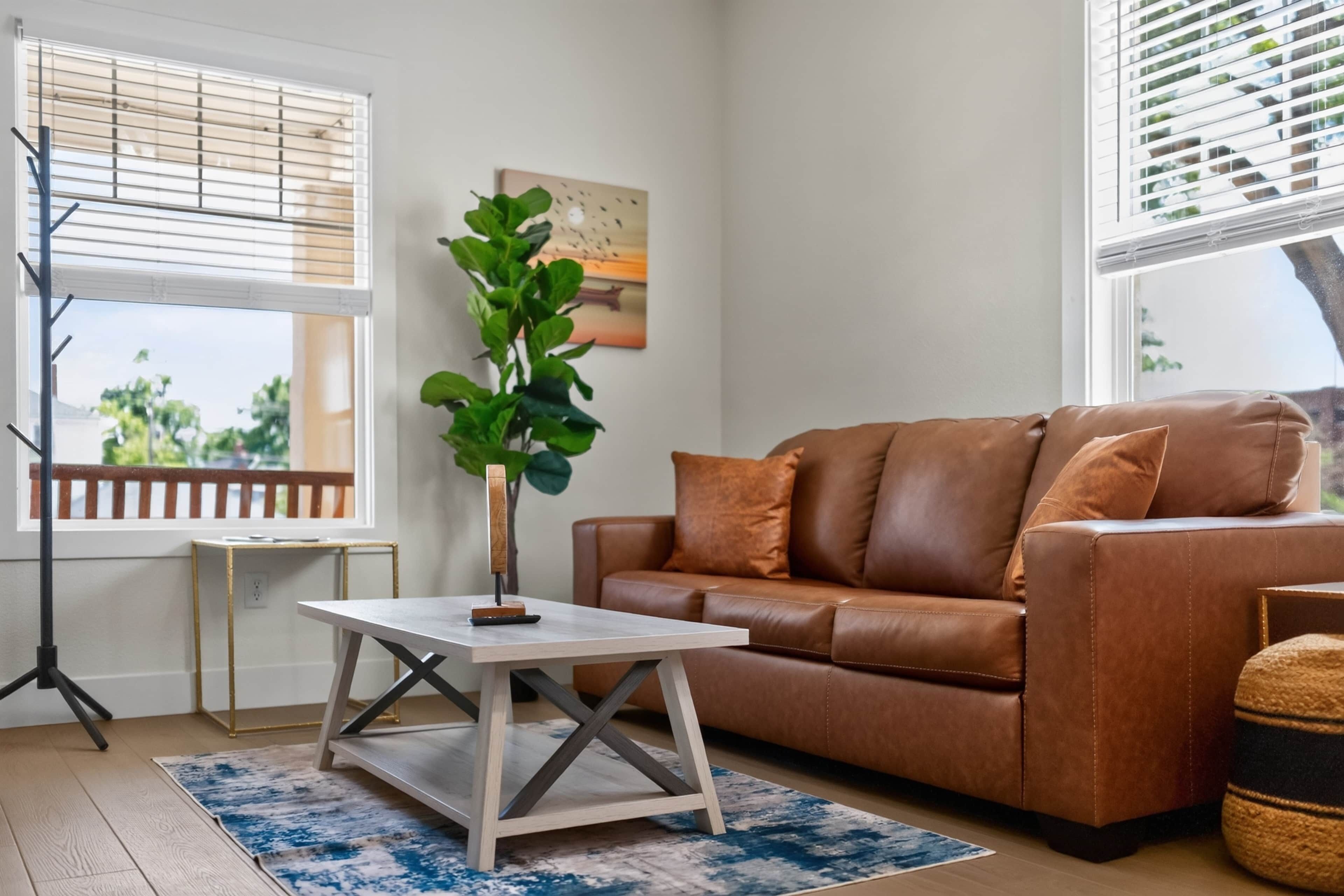 Un salon avec un canapé en cuir marron, une petite table blanche, une plante en pot et une fenêtre avec des stores.