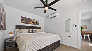 A bedroom with a bed, bedside tables, a ceiling fan, a mounted air conditioner, a painting, and a mirror.