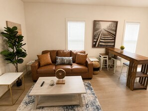 A living room with a brown leather sofa, a coffee table, a dining table, and a potted plant.