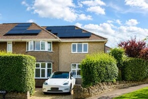 Ein zweigeschossiges Haus mit Solaranlage auf dem Dach, ein weißes Auto auf der Auffahrt und ein gepflegter Heckenstreifen.