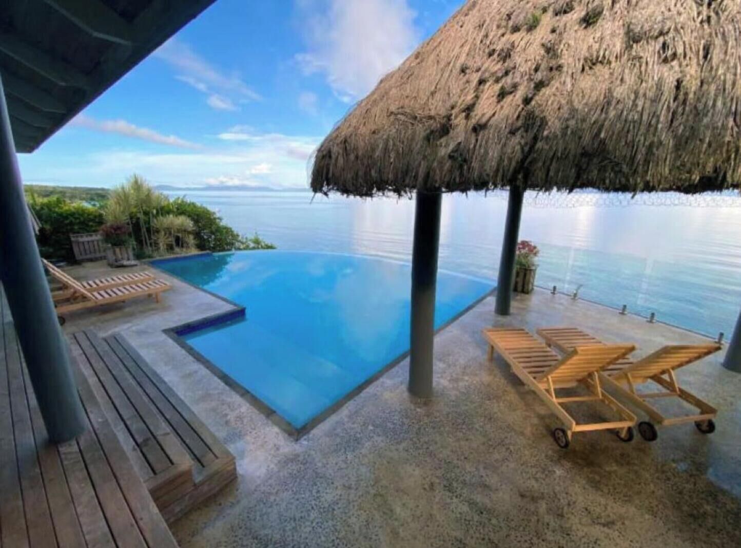 A pool with a thatched roof, wooden lounge chairs, and a view of the sea.