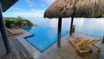 A pool with a thatched roof, wooden lounge chairs, and a view of the sea.