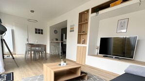 A modern hotel room with a bed, a TV, a wooden coffee table, and a kitchen area in the background.