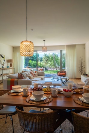 A modern living room with a dining table set for a meal, a sofa with cushions, and a large window offering a view of the outdoors. - Tróia Atlantic Pines Townhouse (Península de Tróia)