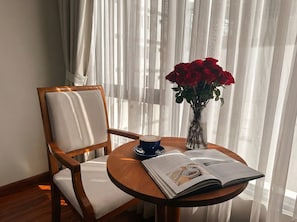 Una silla de madera con cojín, una mesa redonda de madera con un libro y una taza, y un florero con rosas rojas.