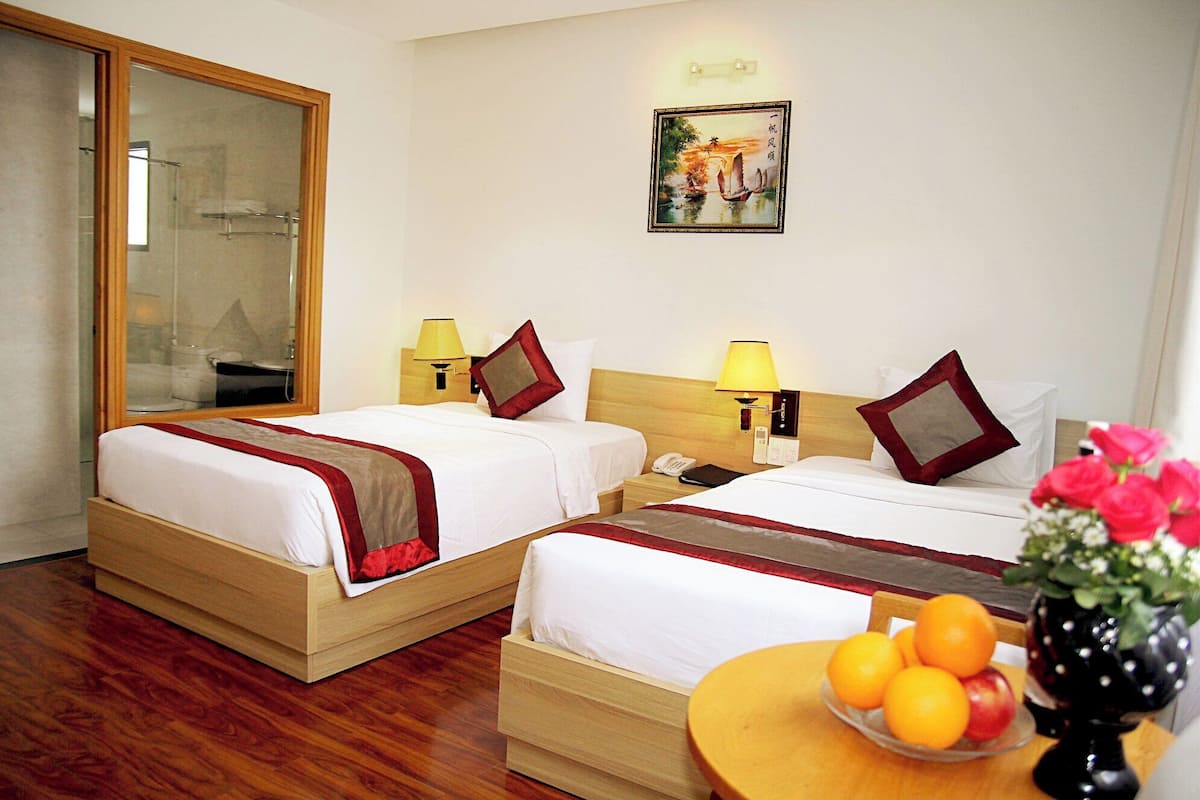 A hotel room with two beds, a wooden floor, a mirror, a painting, a nightstand with a lamp, and a table with fruit.