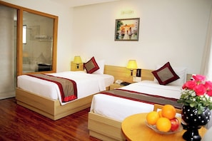 A hotel room with two beds, a wooden floor, a mirror, a painting, a nightstand with a lamp, and a table with fruit.