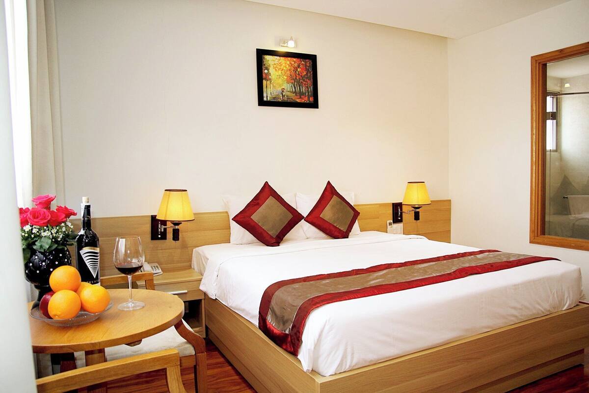 A hotel room with a bed, bedside lamps, a table with fruit and a bottle, and a painting on the wall.