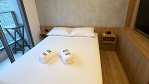 Une chambre d’hôtel avec un lit, une tête de lit en bois, une petite table de chevet en bois, une télévision et une grande fenêtre donnant sur des immeubles.