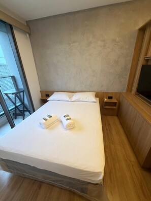 A hotel room with a bed, a wooden headboard, a small wooden nightstand, a TV, and a large window with a view of buildings. - Green Loft - Wonderful City (Rio de Janeiro)