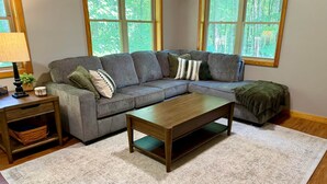 A living room with a sectional sofa, a wooden coffee table, a lamp, and a view of trees through large windows. - Honey Ridge Forest Retreat (Benton Township)