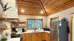 A kitchen with wooden cabinets, a stainless steel refrigerator, a window with a view of trees, and a countertop with various items. - Honey Ridge Forest Retreat (Benton Township)