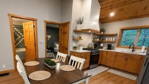 A dining area with a wooden table and chairs, a kitchen with wooden cabinets, and a window with a view of greenery. - Honey Ridge Forest Retreat (Benton Township)