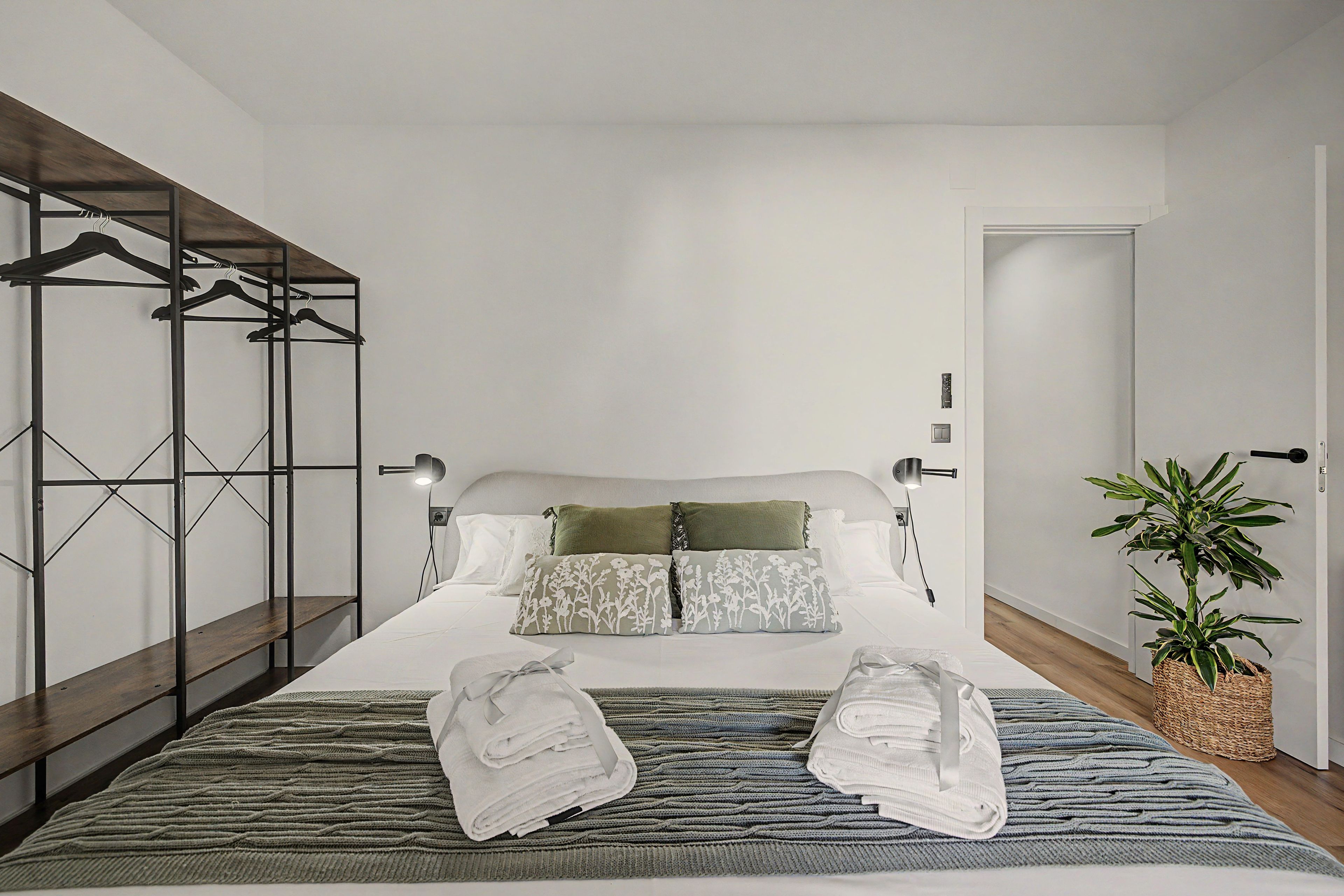 A modern bedroom with a bed, a hanging clothes rack, a bedside lamp, a potted plant, and a folded towel on the bed.