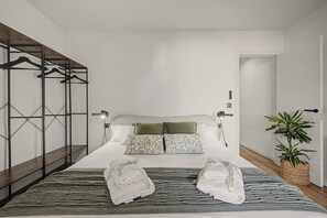 Un dormitorio moderno con cama, perchero, lámpara de noche, planta en maceta y toalla doblada sobre la cama.
