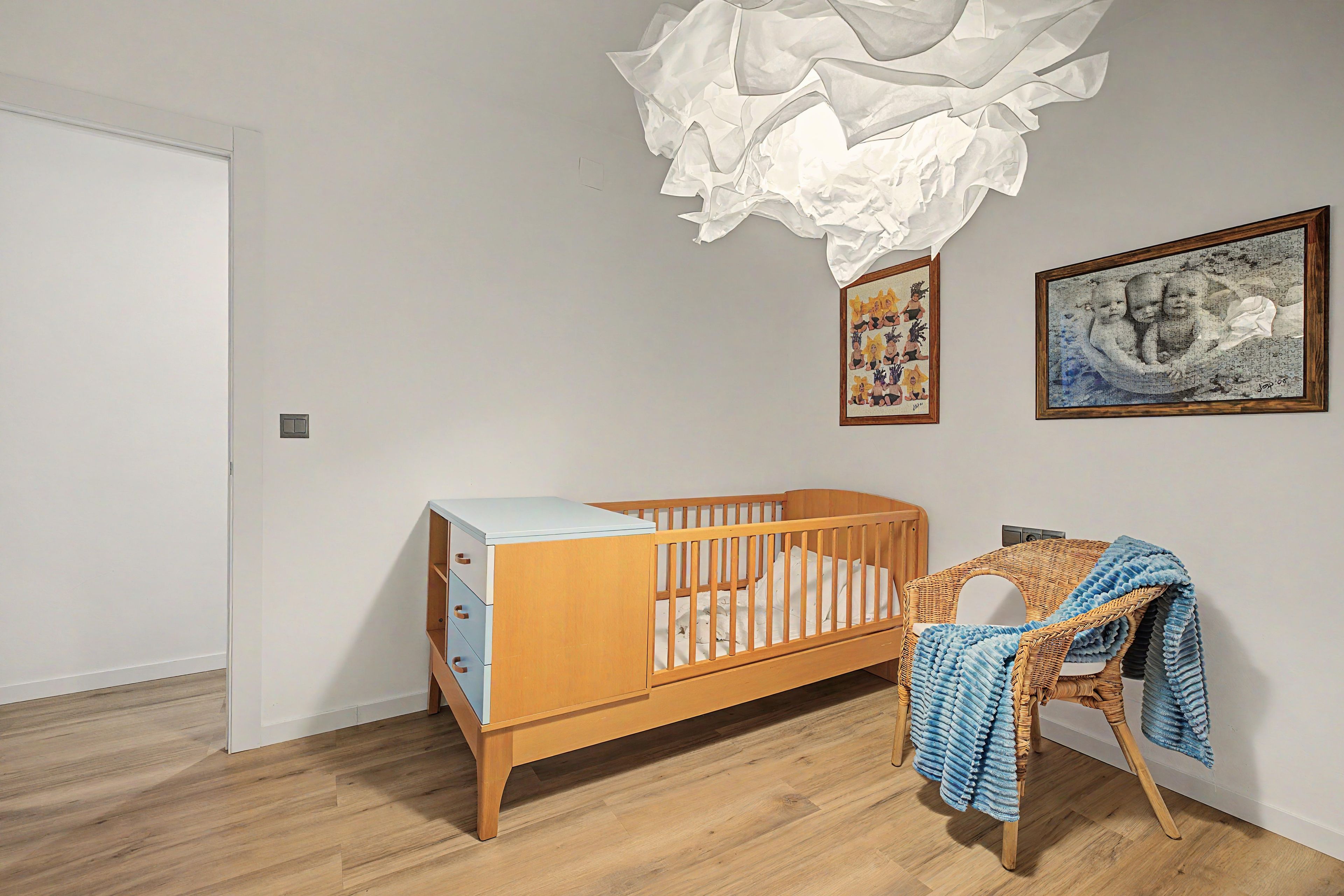 Une pièce avec un berceau en bois, une chaise en osier recouverte d’une couverture bleue et deux œuvres d’art encadrées accrochées au mur.
