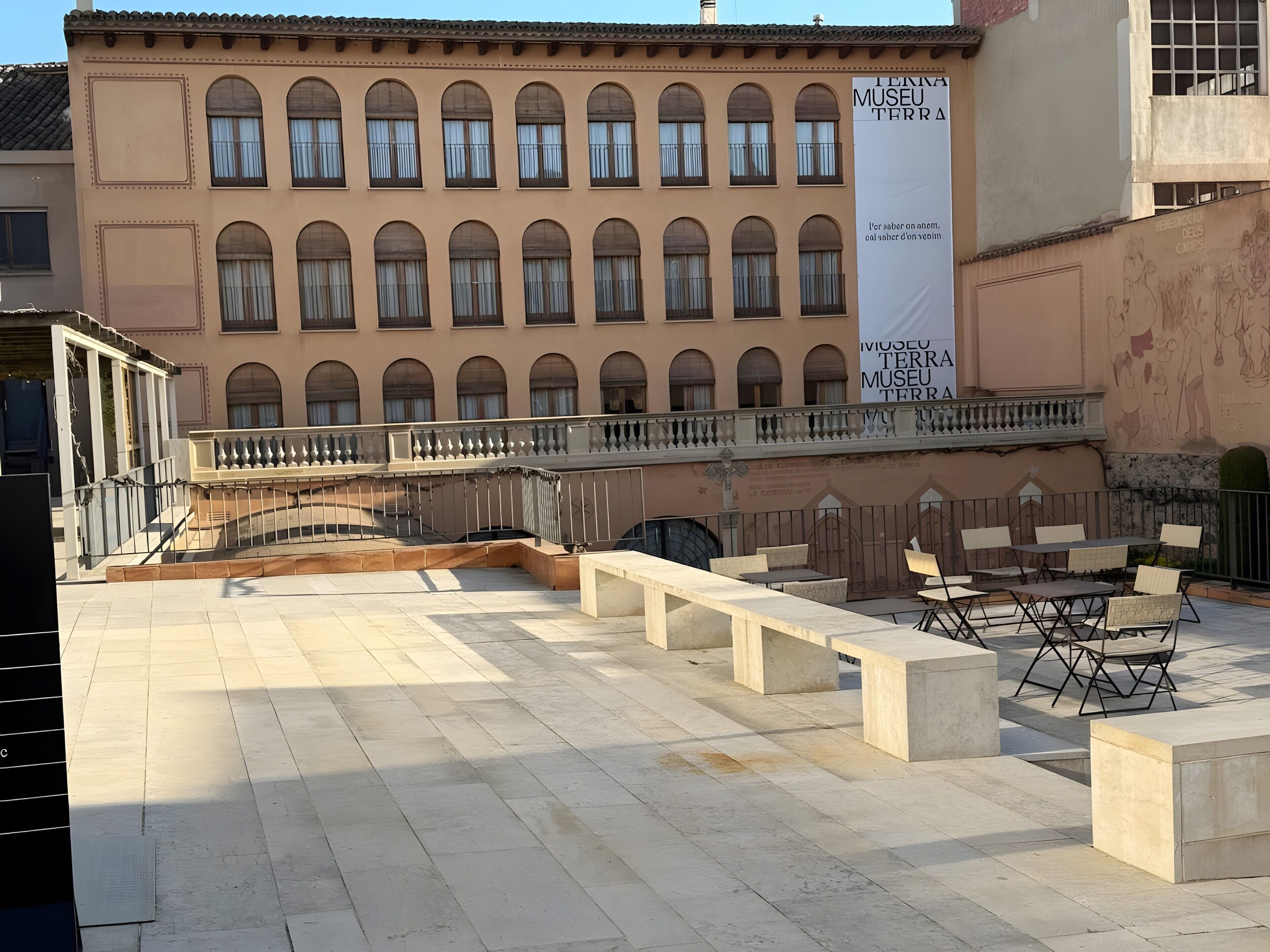 Un patio al aire libre con bancos y mesas de piedra, una barandilla, y un edificio con ventanas de arco y un banderín.